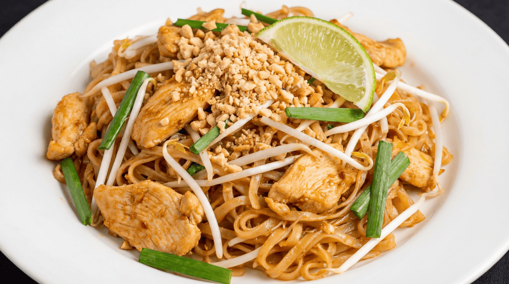 A plate of Chicken Pad Thai topped with chopped peanuts, a lime wedge, bean sprouts, green onions, and tender pieces of chicken, served on a white dish.
