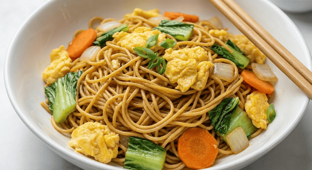 A bowl of Chinese Egg Stir Fry Noodles with scrambled eggs, baby bok choy, sliced carrots, onions, and chopped green onions, served with wooden chopsticks on the edge of the bowl.